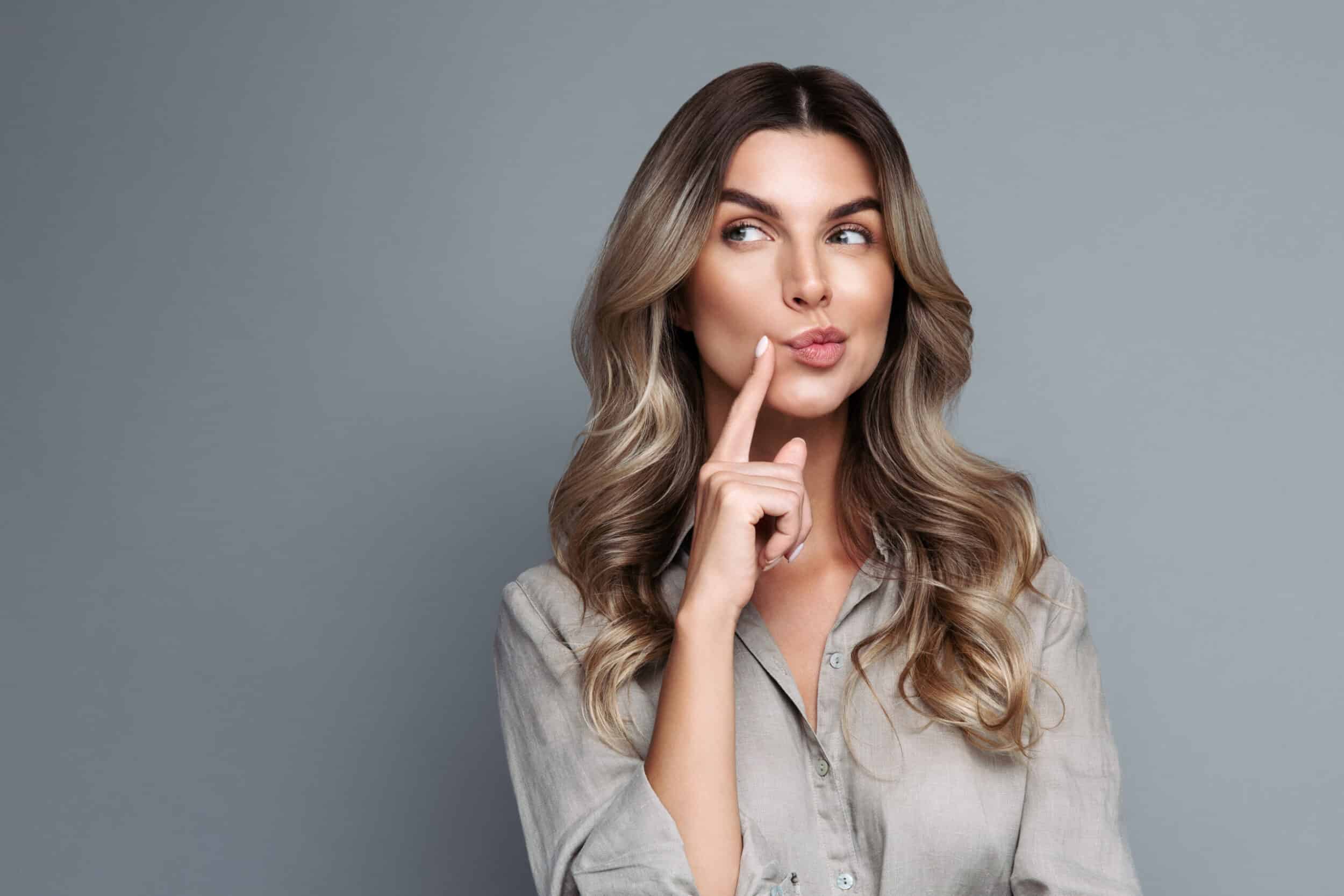 Serious young blonde woman with curly hair dressed in linen shirt thinking, having tricky plan in mind, looking away at blank space for your advertisement, isolated on studio grey background.