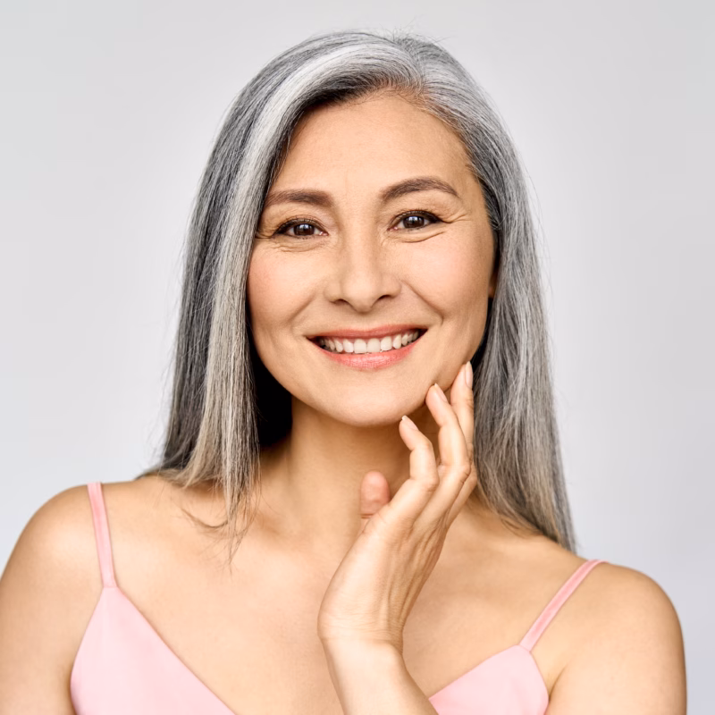 Headshot of a happy, mature woman in her 50s touching her face and smiling at the camera, isolated on white, representing anti-wrinkle skincare, lifting treatments, and Fractional CO2 therapy